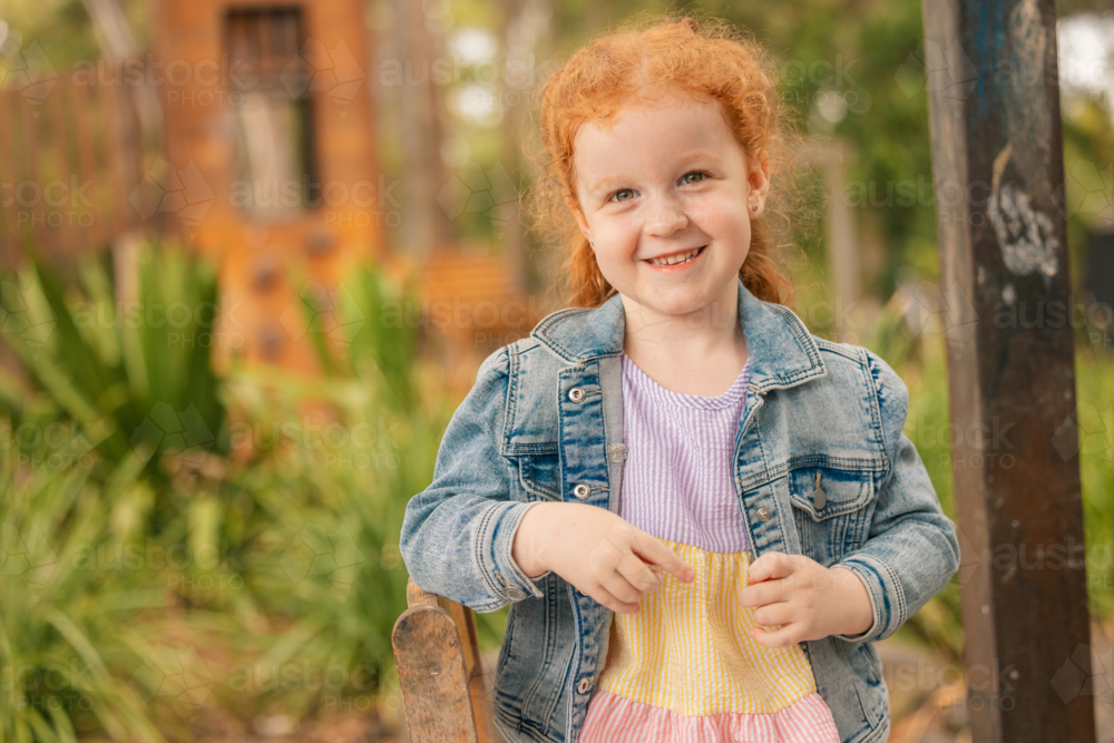 Outdoor portrait of happy preschool age girl with long red hair at kindergarten - Australian Stock Image