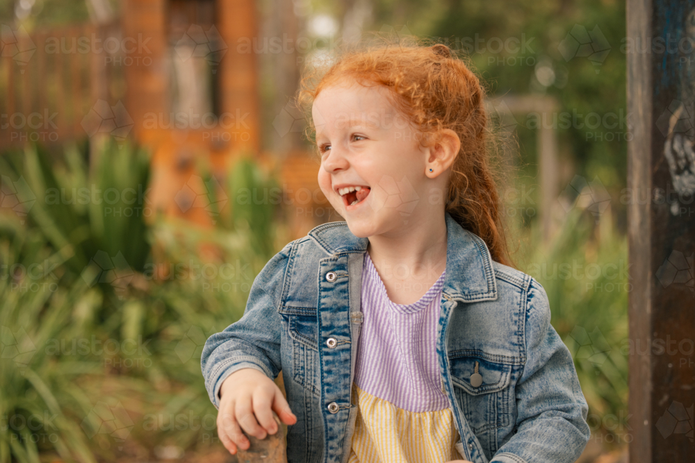 Outdoor portrait of happy preschool age girl with long red hair looking away with big smile - Australian Stock Image