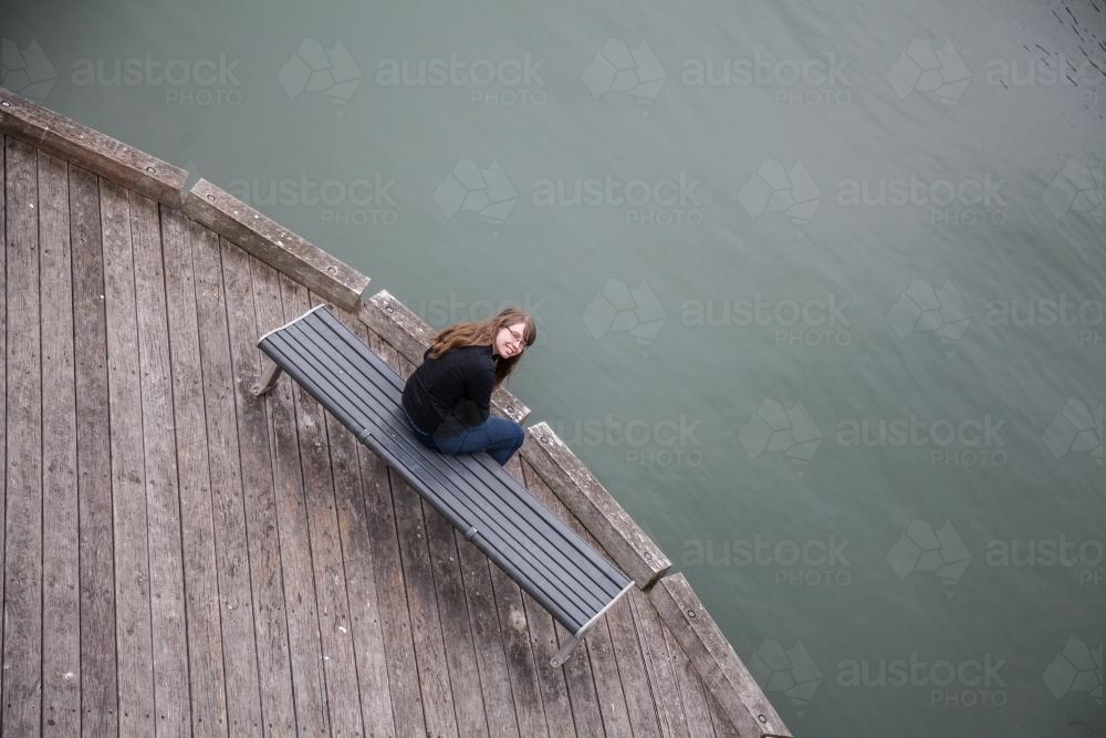 Over head view of teen girl sitting on wooden seat beside water - Australian Stock Image