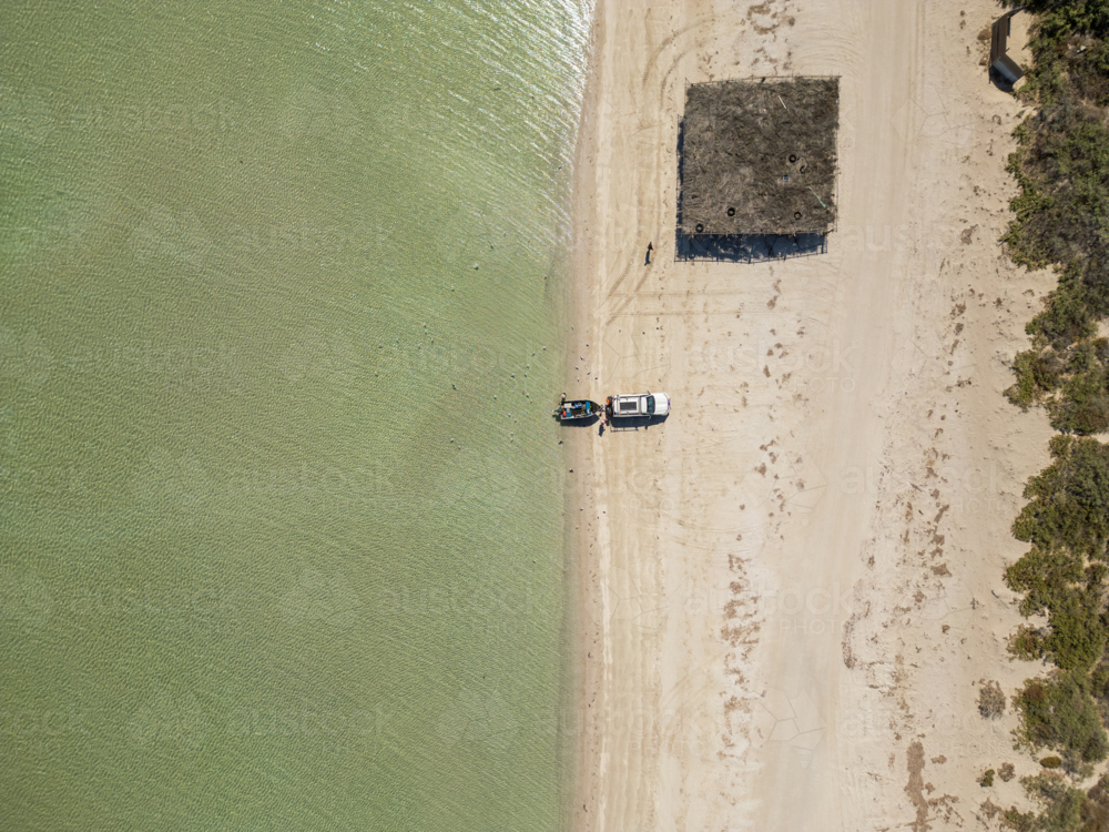 Overhead view of a  white vehicle and small boat sit at the edge of a sandy shoreline - Australian Stock Image