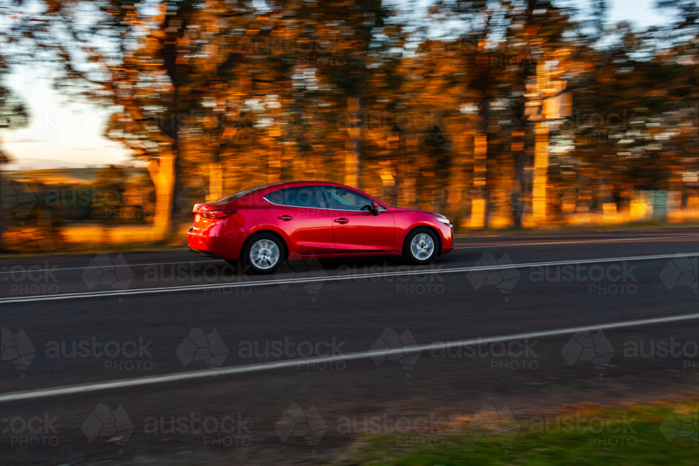 Panning image of red car driving past on sealed rural country road in sunset light - Australian Stock Image