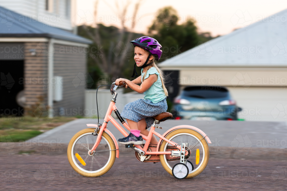 Panning movement shot of preschooler riding pink bike with training wheels on roadside - Australian Stock Image