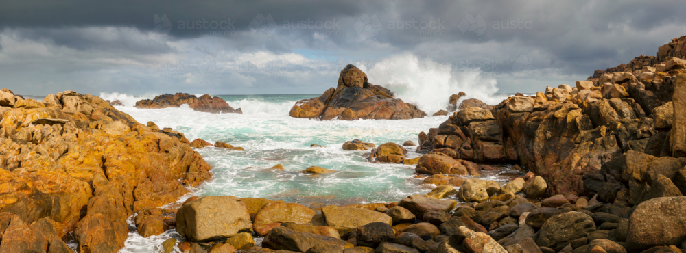 Panorama view of large ocean waves crashing over coastal rock formations under a dark stormy sky - Australian Stock Image