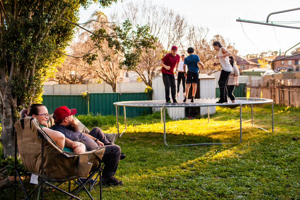 Parents watching australian kids jumping on a trampoline without an enclosure in backyard - Australian Stock Image