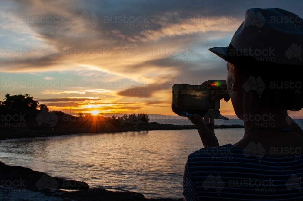 Partial silhouette of woman taking photo of sunset on mobile phone - Australian Stock Image