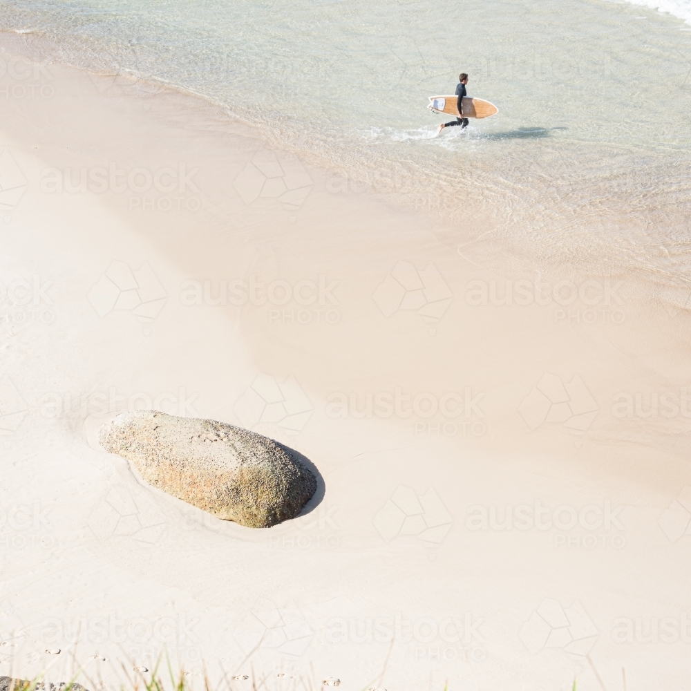 pastel tone beach with lone rock and surfer walking into sea - Australian Stock Image