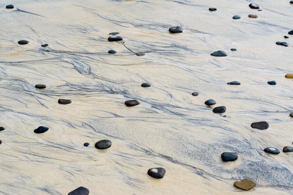 Patterns in wet yellow and black mineral sands on a beach at low tide with pebbles - Australian Stock Image
