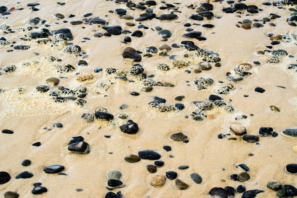 Pebbles scattered on a sand beach at low tide with foam - Australian Stock Image