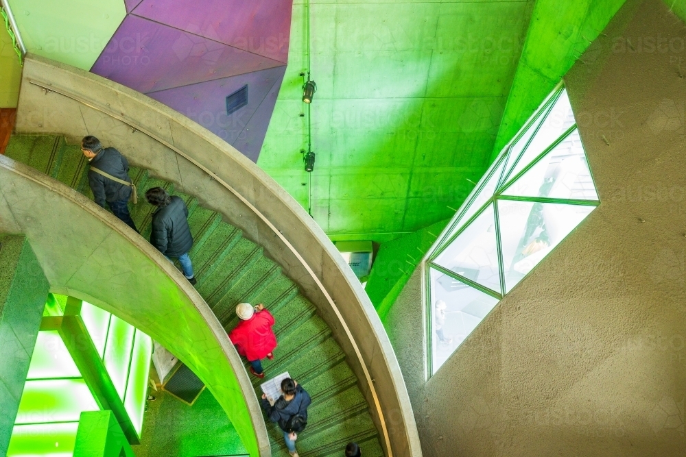 People climbing a curved staircase in a brightly coloured building interior. - Australian Stock Image