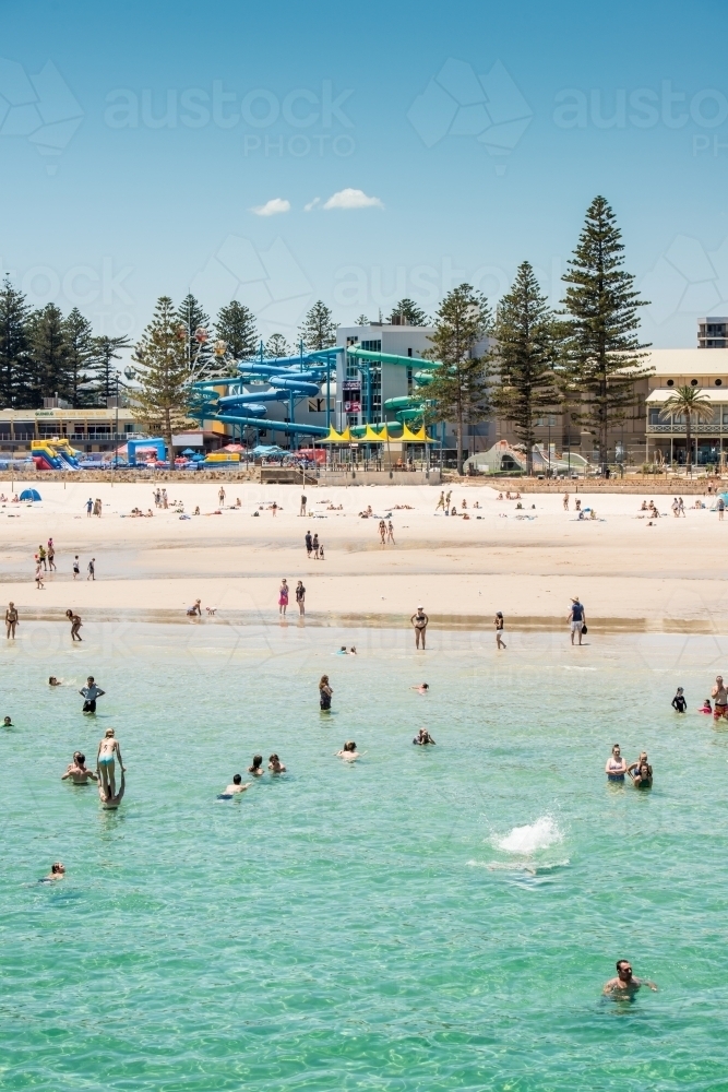 People in the water and on the sand at the beach, with waterslide in the background - Australian Stock Image