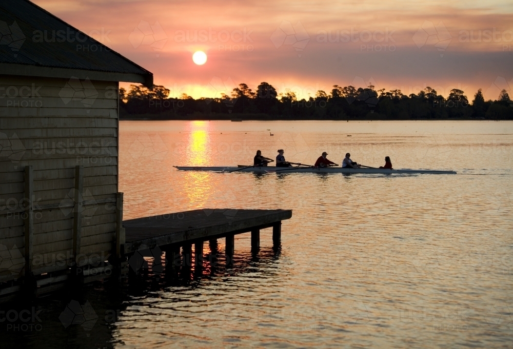 People rowing past a jetty on a lake at sunset - Australian Stock Image