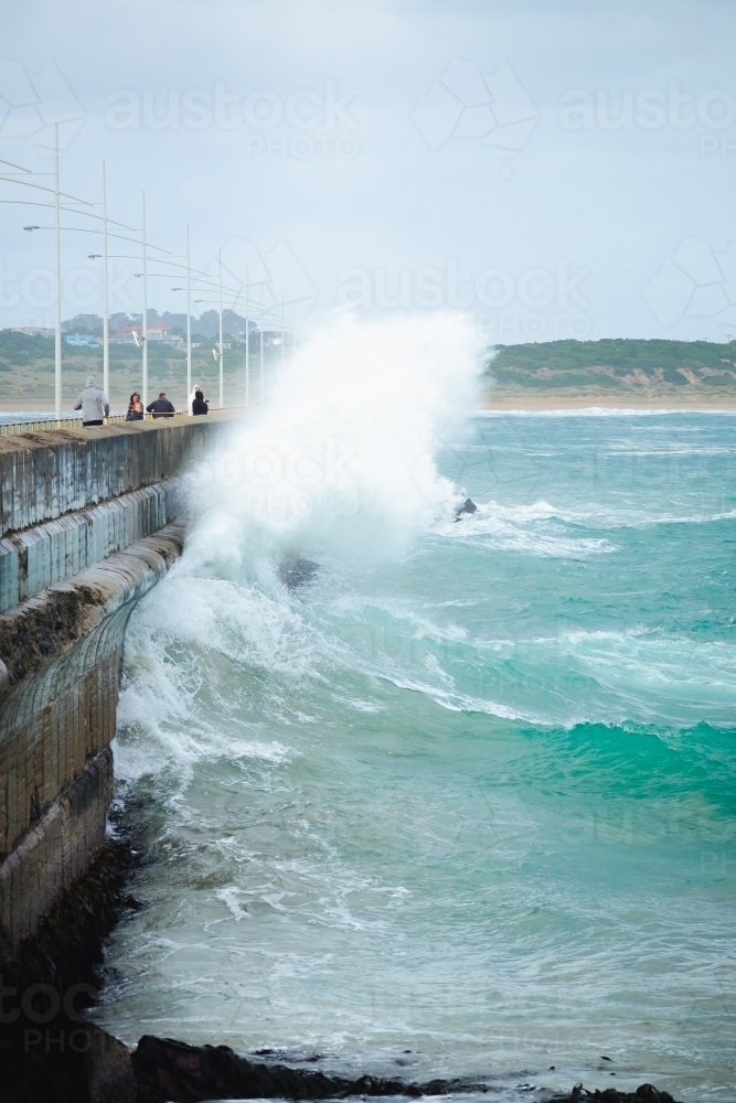 People walk along the breakwater as a huge wave crashes - Australian Stock Image