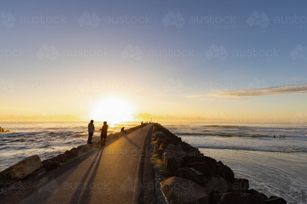 People walking along rock wall at sunrise with the ocean waves breaking on both sides - Australian Stock Image
