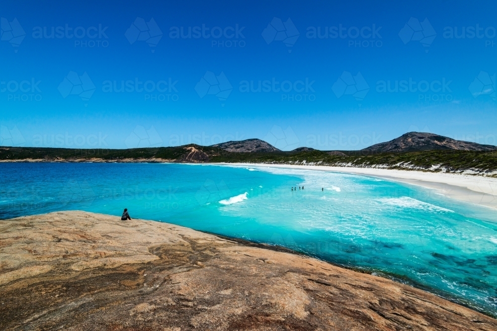 Person sitting looking over white sandy beach with people swimming in aqua blue water - Australian Stock Image
