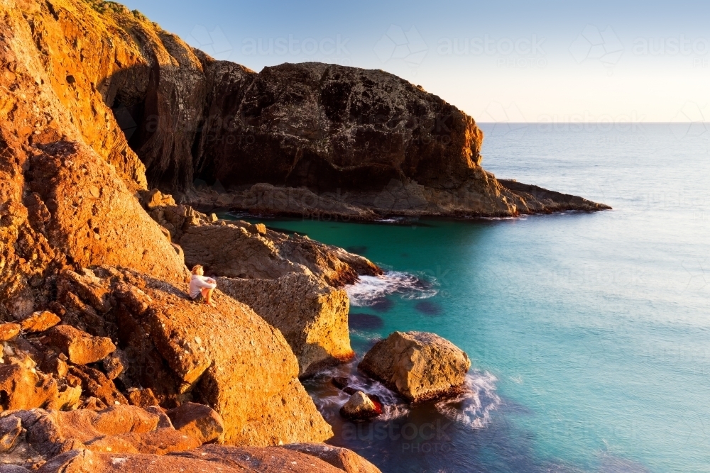 Person sitting on a sunlit cliff edge watching the ocean - Australian Stock Image