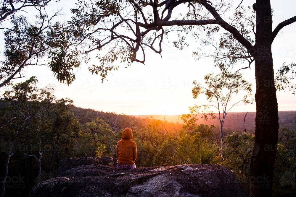 Person sitting on remote rock on hiking trail overlooking sunrise - Australian Stock Image
