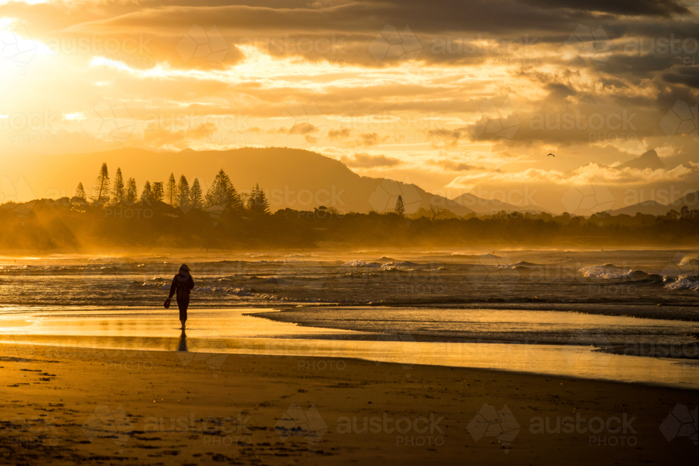Person walking along a beach at sunset with golden light and distant mountains - Australian Stock Image