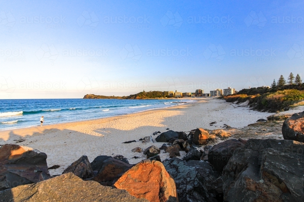 Person walking along sandy beach and rocky coastline beneath blue sky - Australian Stock Image
