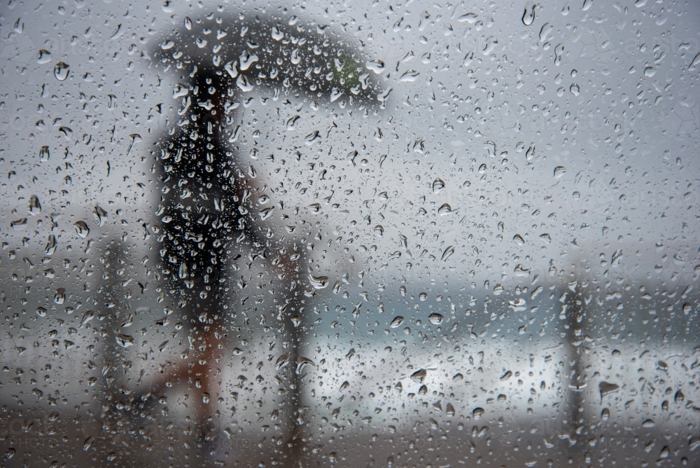 Person walking in rain with an umbrella, shot through car window with drops on glass - Australian Stock Image