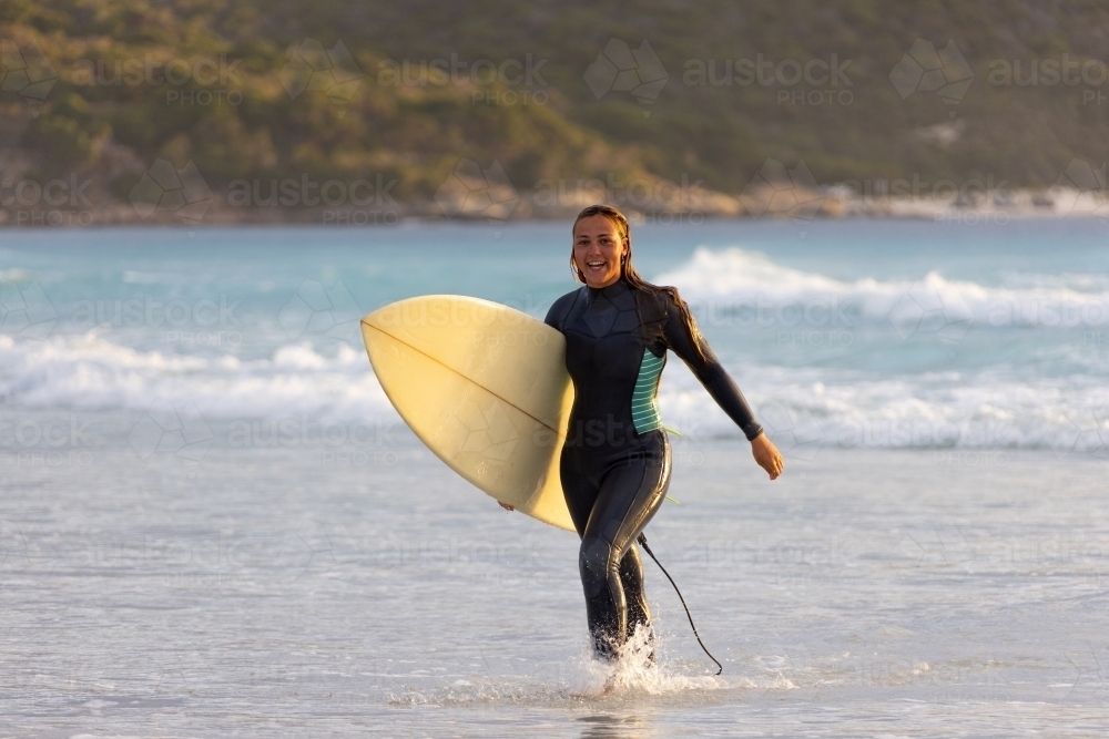 Person walking out of the water carrying surfboard on beach - Australian Stock Image