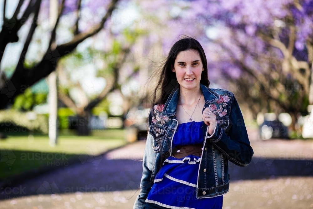 Portrait of young Australian person walking outside in spring smiling with jacaranda trees overhead - Australian Stock Image