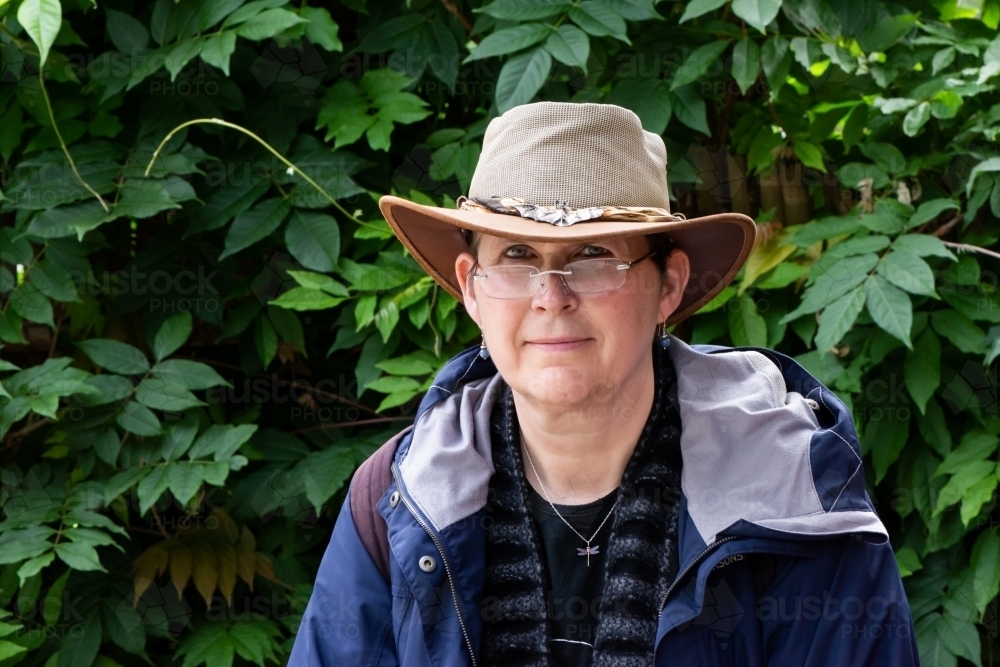 Portrait of a female tourist wearing an Australian hat, glasses and rain jacket with greenery behind - Australian Stock Image
