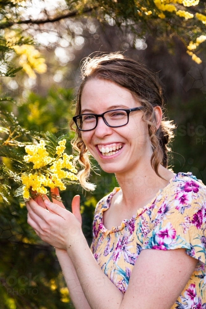 Portrait of a happy laughing young woman with glasses standing among leaves and blossoms of wattle - Australian Stock Image