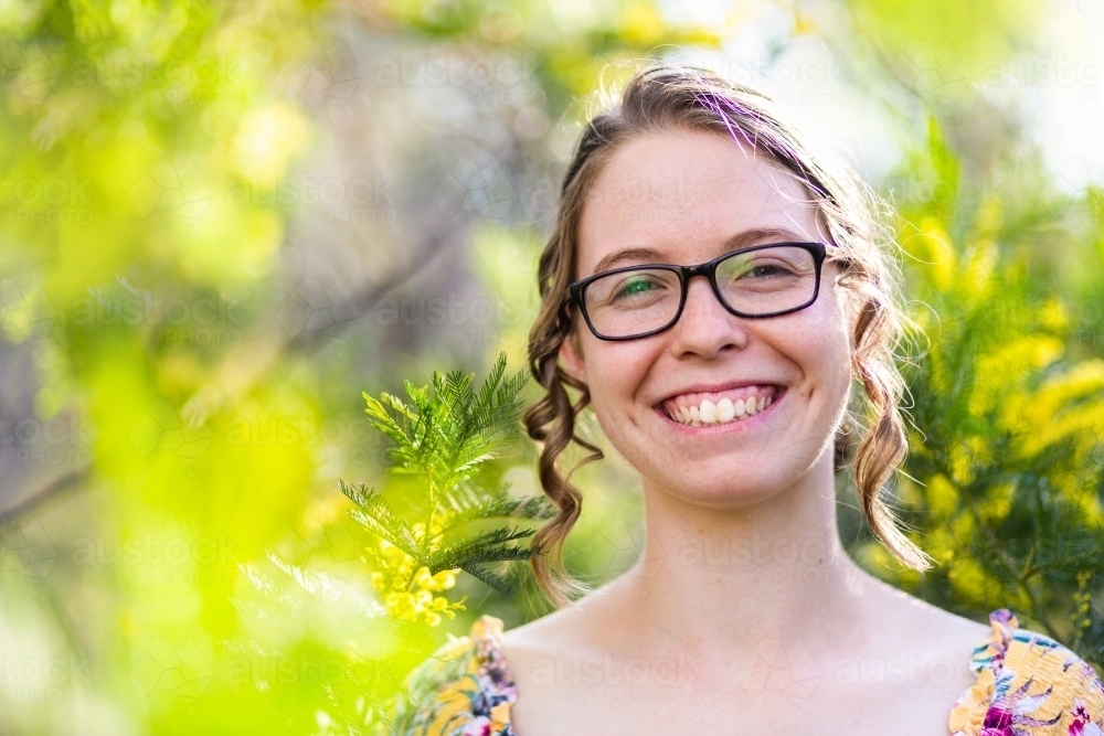 Portrait of a happy smiling young woman with glasses standing among leaves and blossoms of wattle - Australian Stock Image