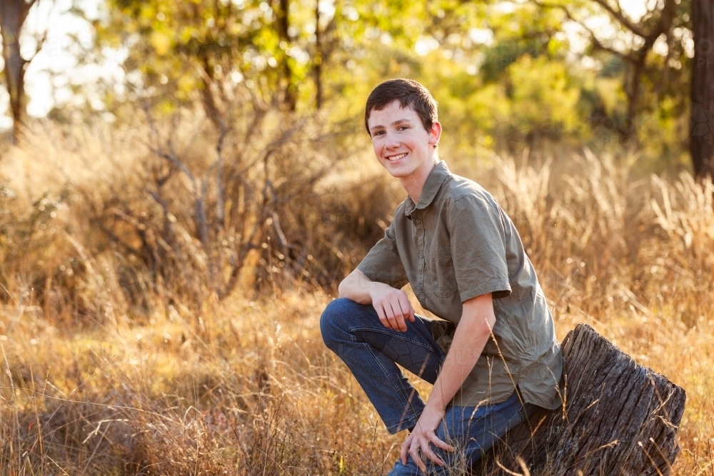 Portrait of a happy teen boy leaning back on tree stump in australian paddock with copy space - Australian Stock Image