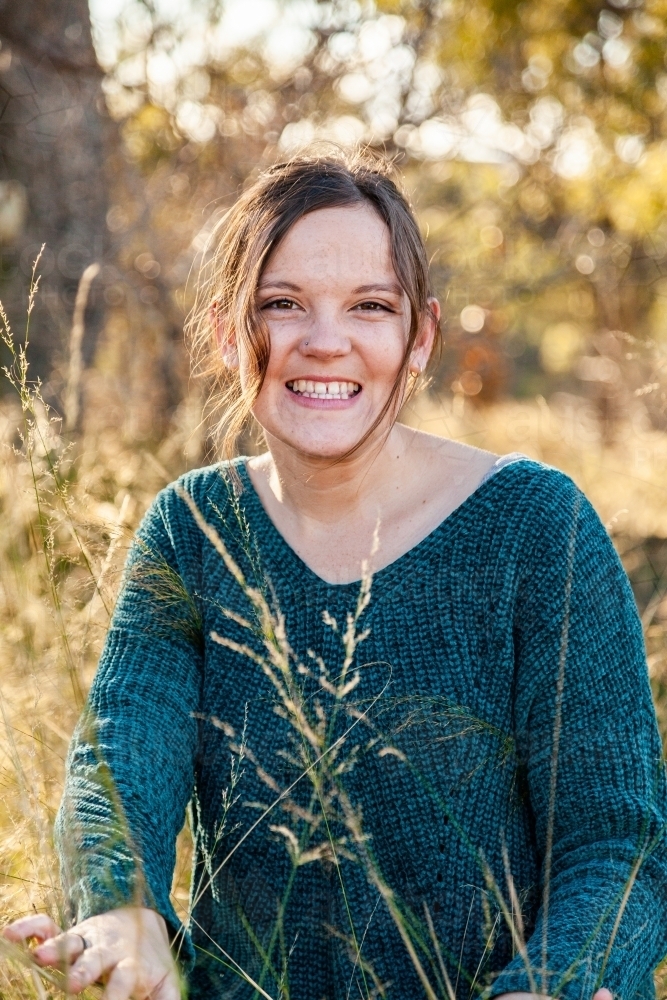Portrait of a happy young adult in green jumper siting in backlit grass - Australian Stock Image