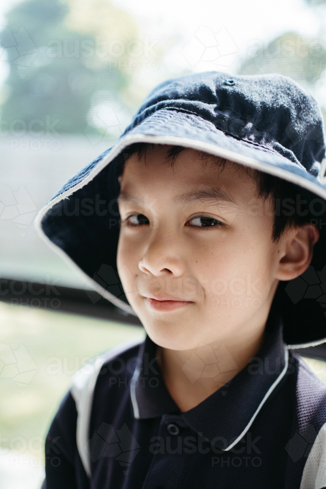 Portrait of a happy young boy in school uniform wearing a bucket hat indoors - Australian Stock Image