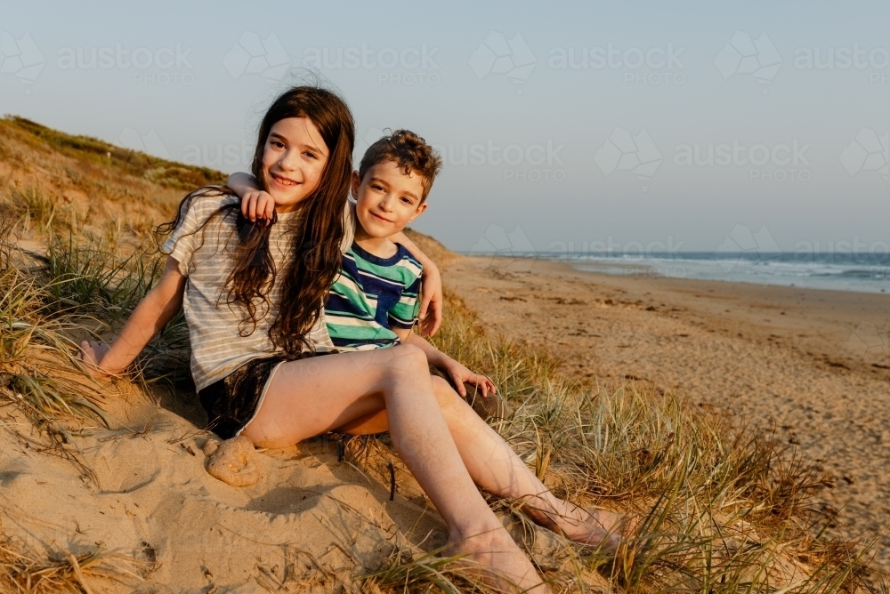 Portrait of a happy young brother & sister sitting on a sand dune; Thirteenth Beach, Barwon Heads - Australian Stock Image