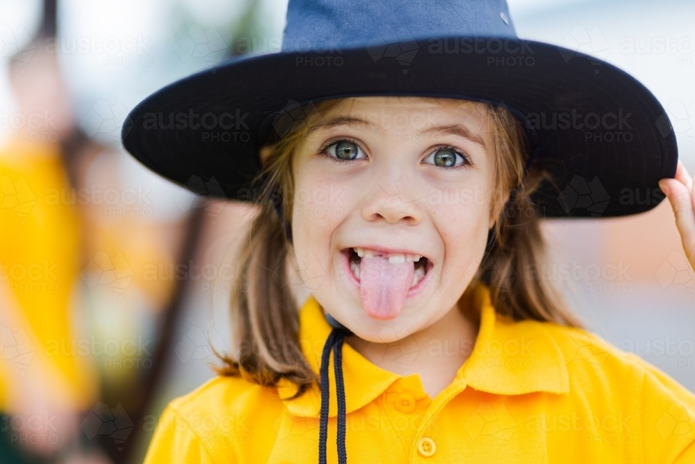 Portrait of a happy young school girl making a silly face outside wearing a hat for sun protection - Australian Stock Image