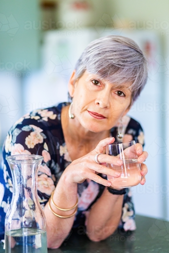 Portrait of a healthy senior woman leaning on the kitchen bench drinking a glass of water - Australian Stock Image
