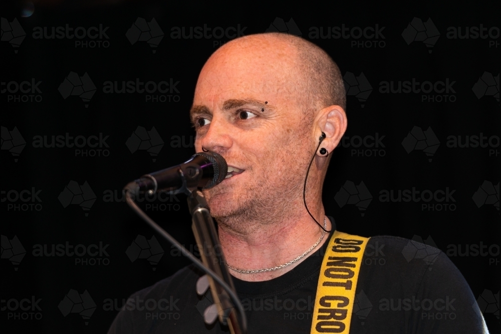 Portrait of a male entertainer singing at microphone with shaved head and black background - Australian Stock Image