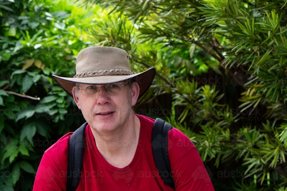 Portrait of a mature male tourist wearing an Australian hat and a backpack with greenery behind - Australian Stock Image
