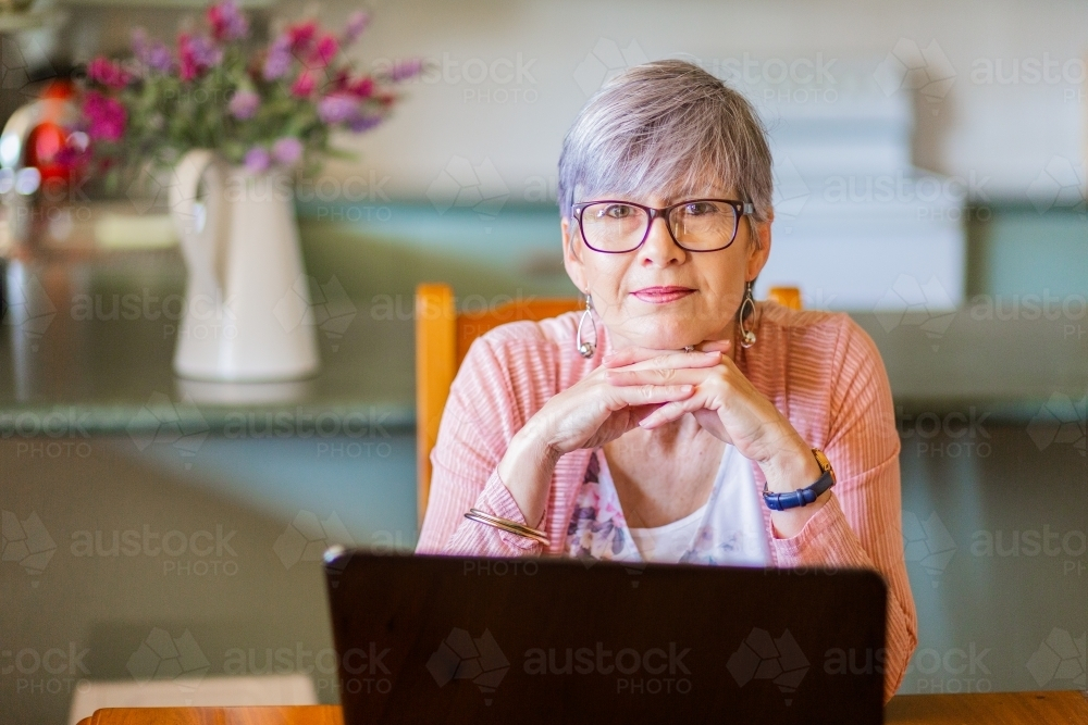 Portrait of a senior woman wearing glasses with hands under her chin sitting inside her home - Australian Stock Image