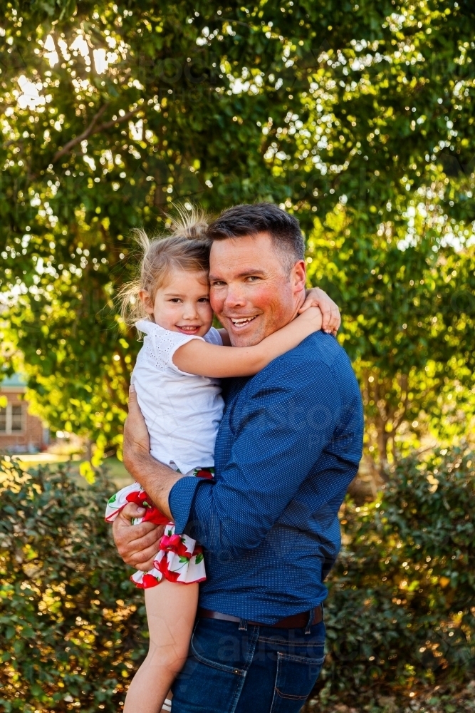 Portrait of a smiling father hugging his happy daughter in golden sunlight - Australian Stock Image