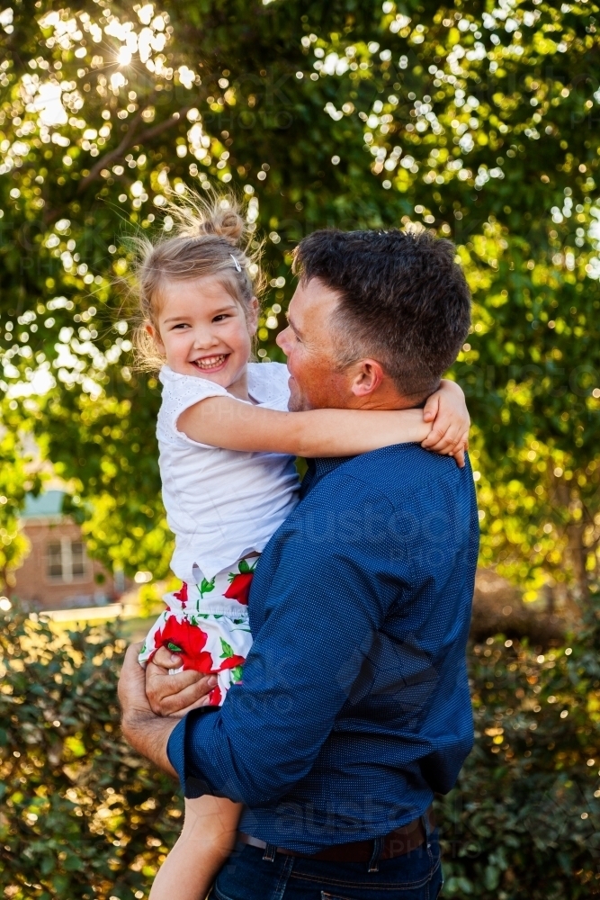 Portrait of a smiling father hugging his happy daughter in golden sunlight - Australian Stock Image