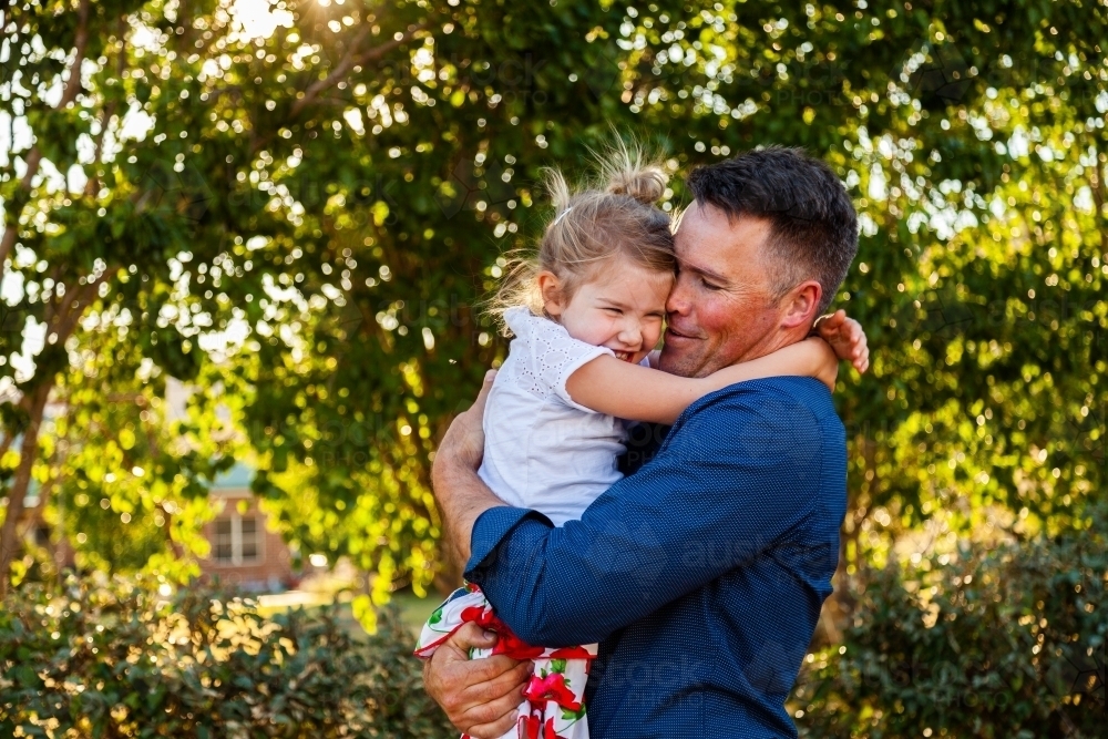 Portrait of a smiling father hugging his happy daughter in golden sunlight - Australian Stock Image