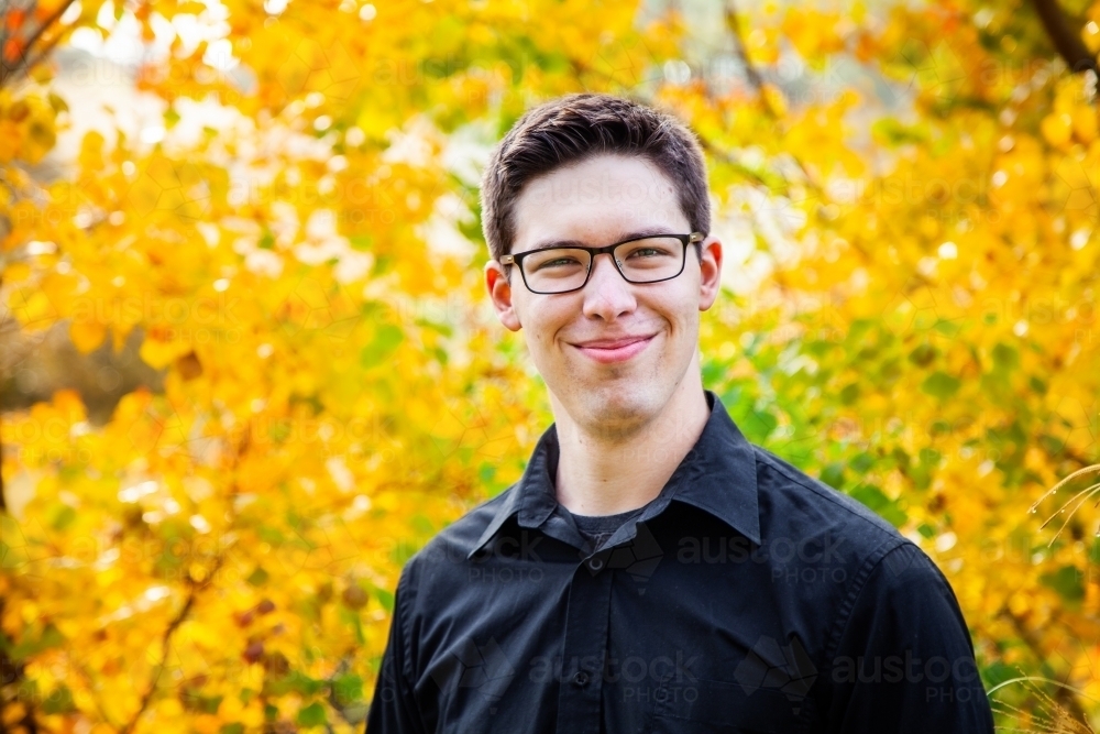 Portrait of a smiling young man in black button up with background of autumn leaves - Australian Stock Image