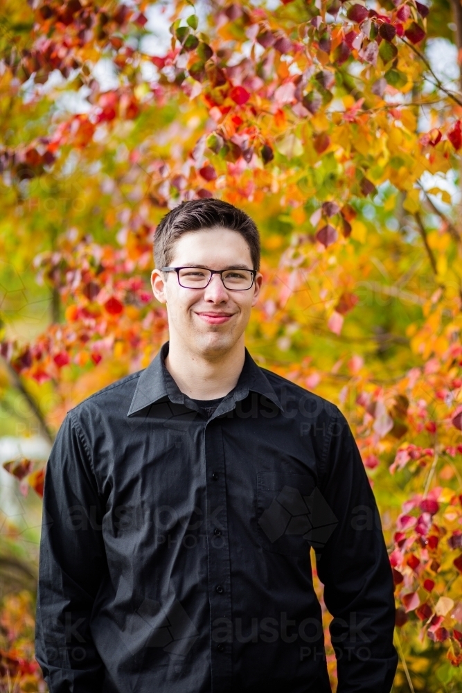 Portrait of a smiling young man in black button up with background of autumn leaves - Australian Stock Image