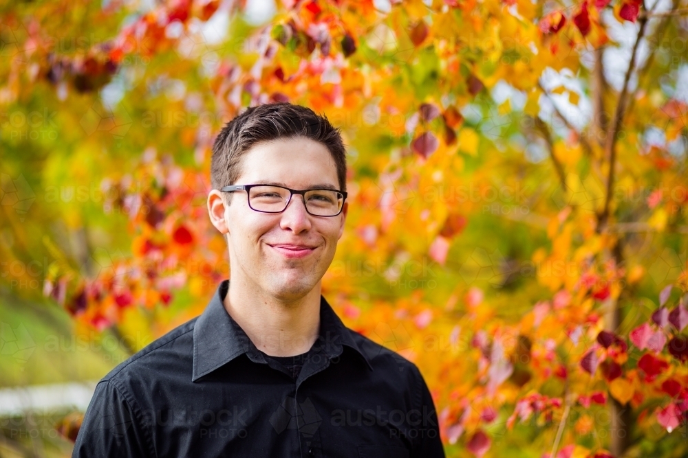 Portrait of a smiling young man in black button up with background of autumn leaves - Australian Stock Image