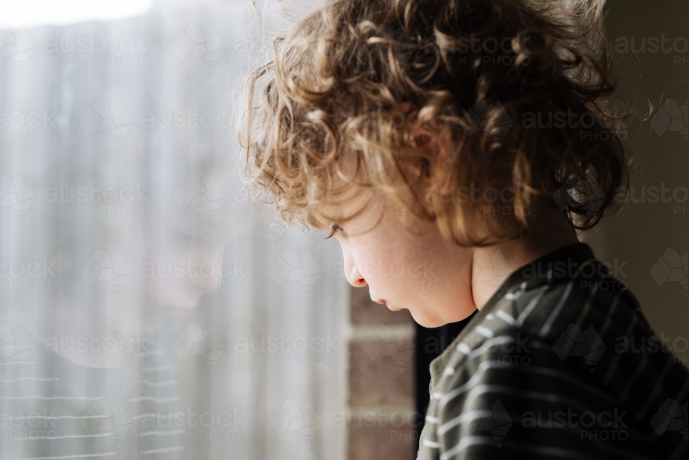Portrait of a young boy leaning his head against a window alone looking sad - Australian Stock Image