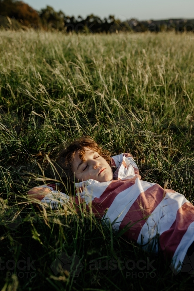 Portrait of a young boy lying with his hands behind his head in a tall grass field at sunset - Australian Stock Image