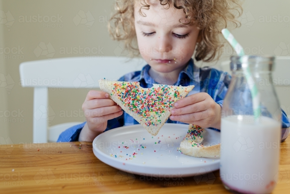 Portrait of a young boy with blond curly hair sitting at the table eating fairy bread - Australian Stock Image