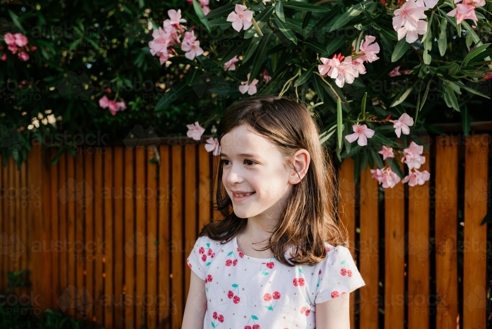 Portrait of a young girl outdoors in front of a wooden fence and garden with pink flowers - Australian Stock Image