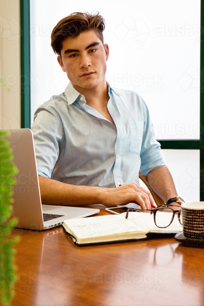 Portrait of a young male office worker sitting with laptop and note book at a meeting table - Australian Stock Image
