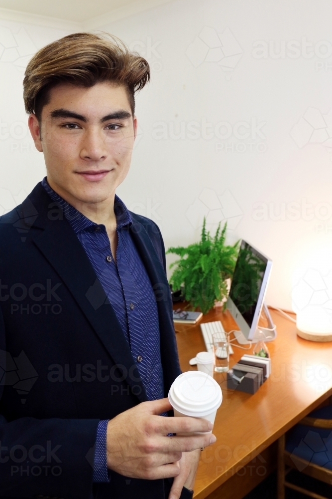 Portrait of a young professional office worker standing with a coffee in front of his desk - Australian Stock Image