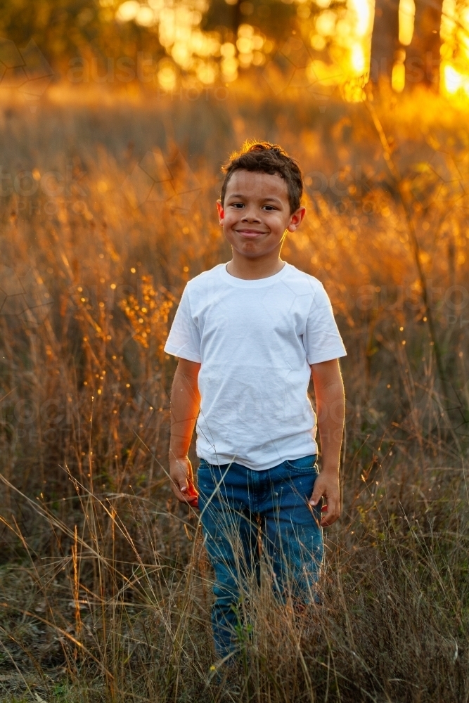 Portrait of aboriginal kid standing in grassy paddock at sunset - Australian Stock Image